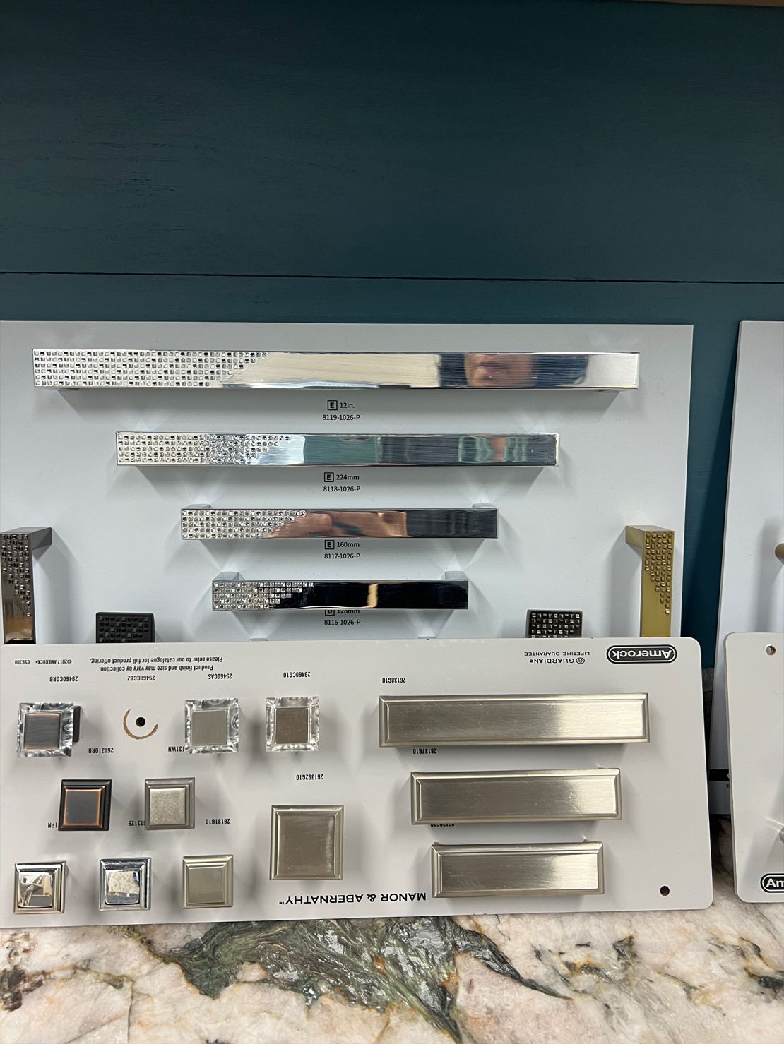 Display of cabinet hardware samples in various metallic finishes against a teal background.