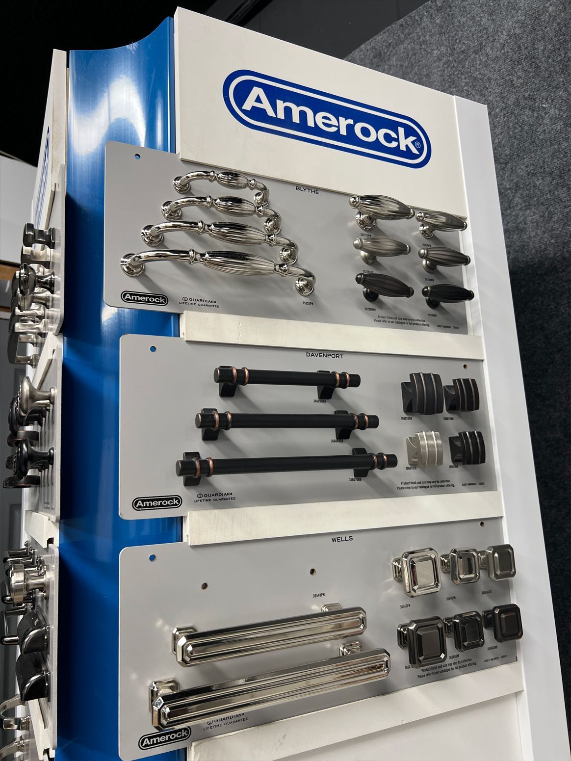 Amerock cabinet hardware display with silver, black, and gold handles on white and blue stand.