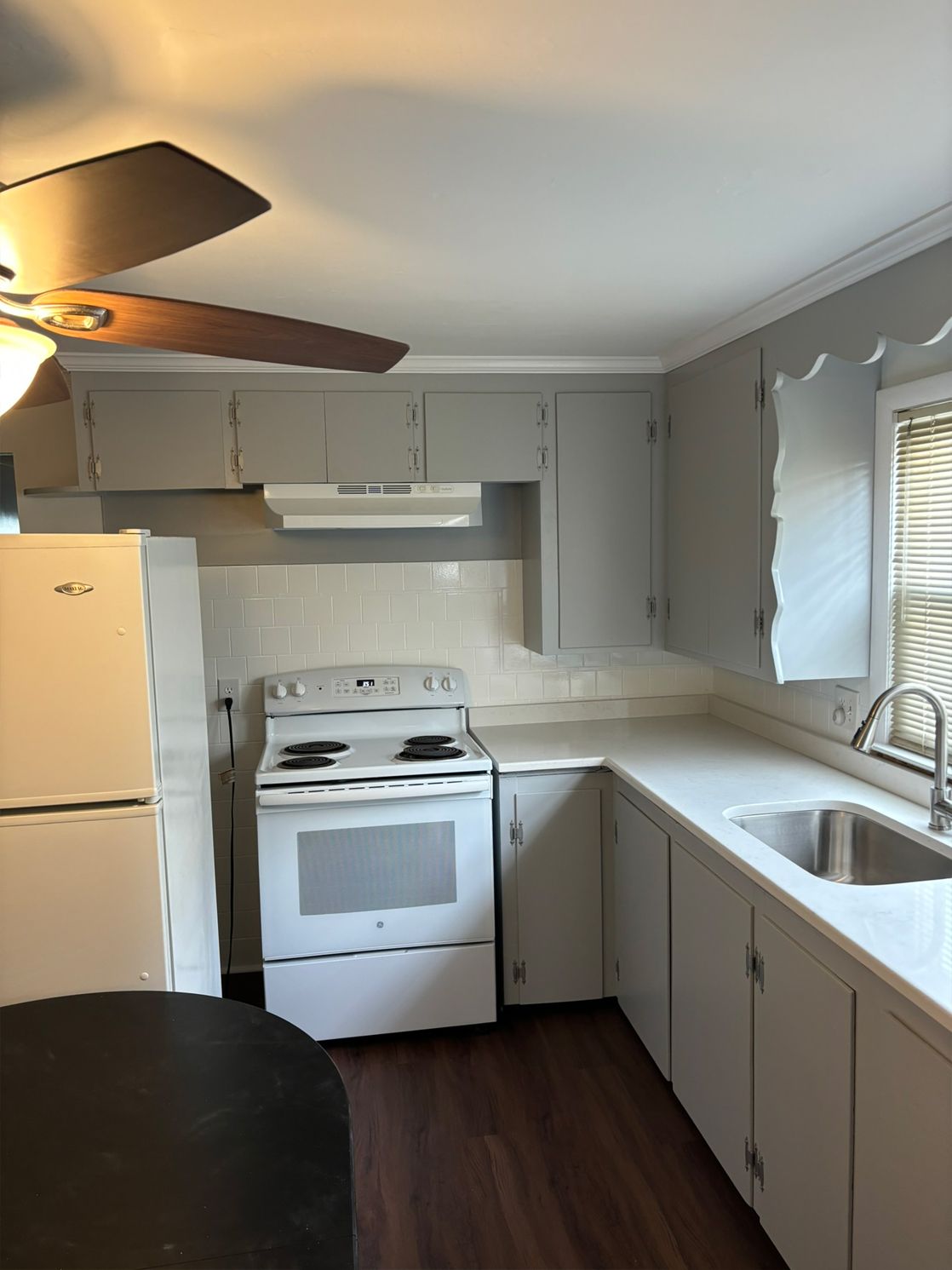 A small kitchen with white appliances and cabinets, a stainless steel sink, and a dark brown floor.