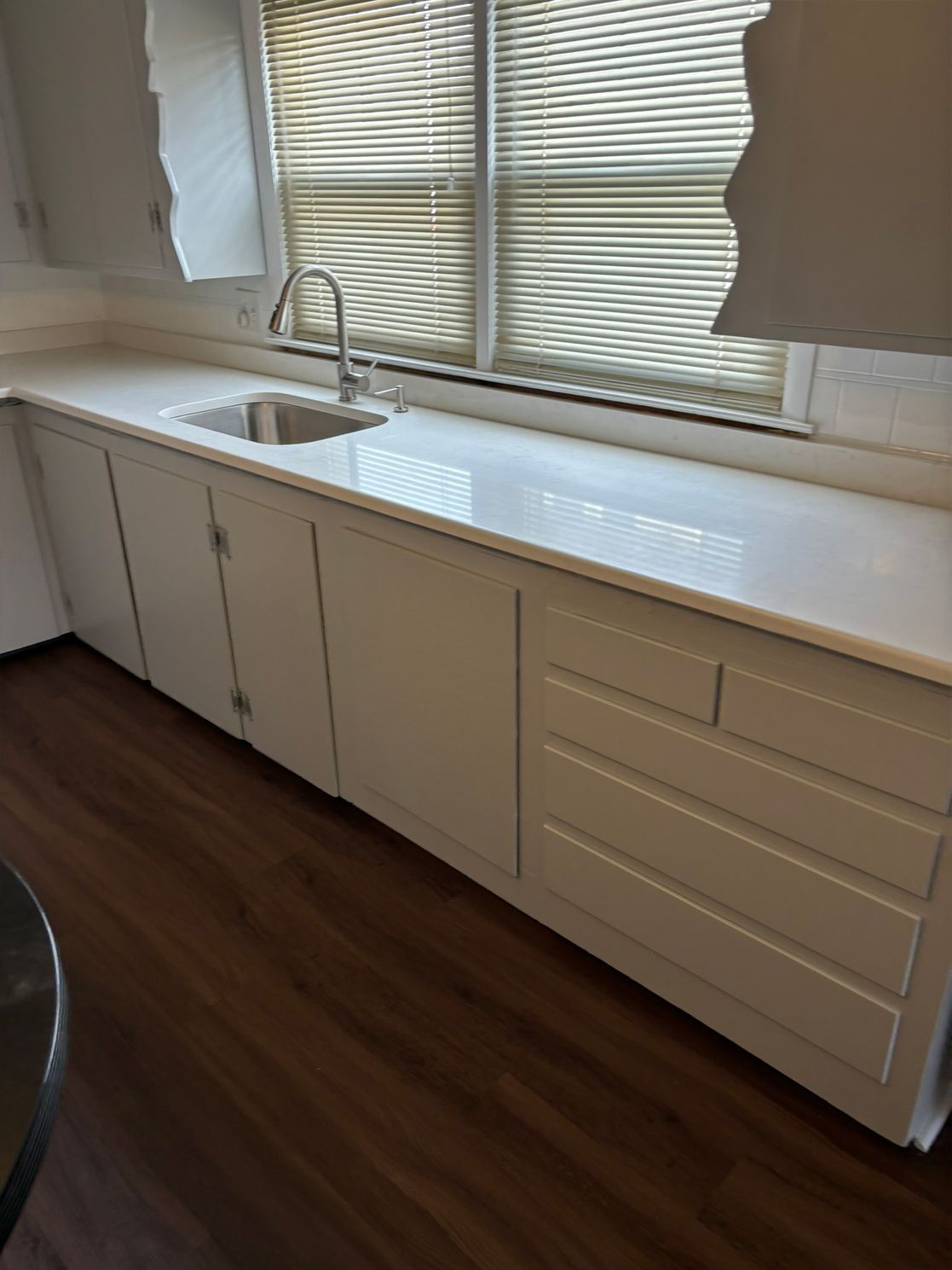 White kitchen cabinets with a white countertop and a window above the sink. Dark wood flooring.