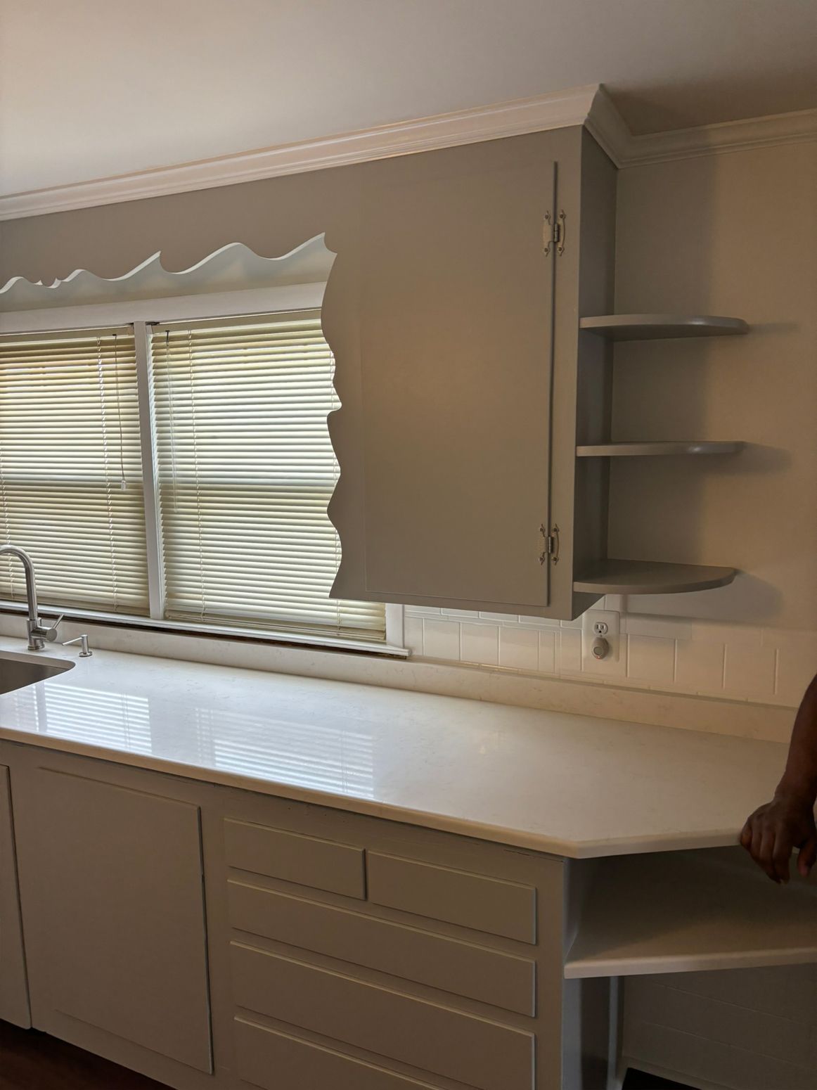 Kitchen with light-colored cabinets, white countertop, and window with blinds. Open cabinet on the right side.