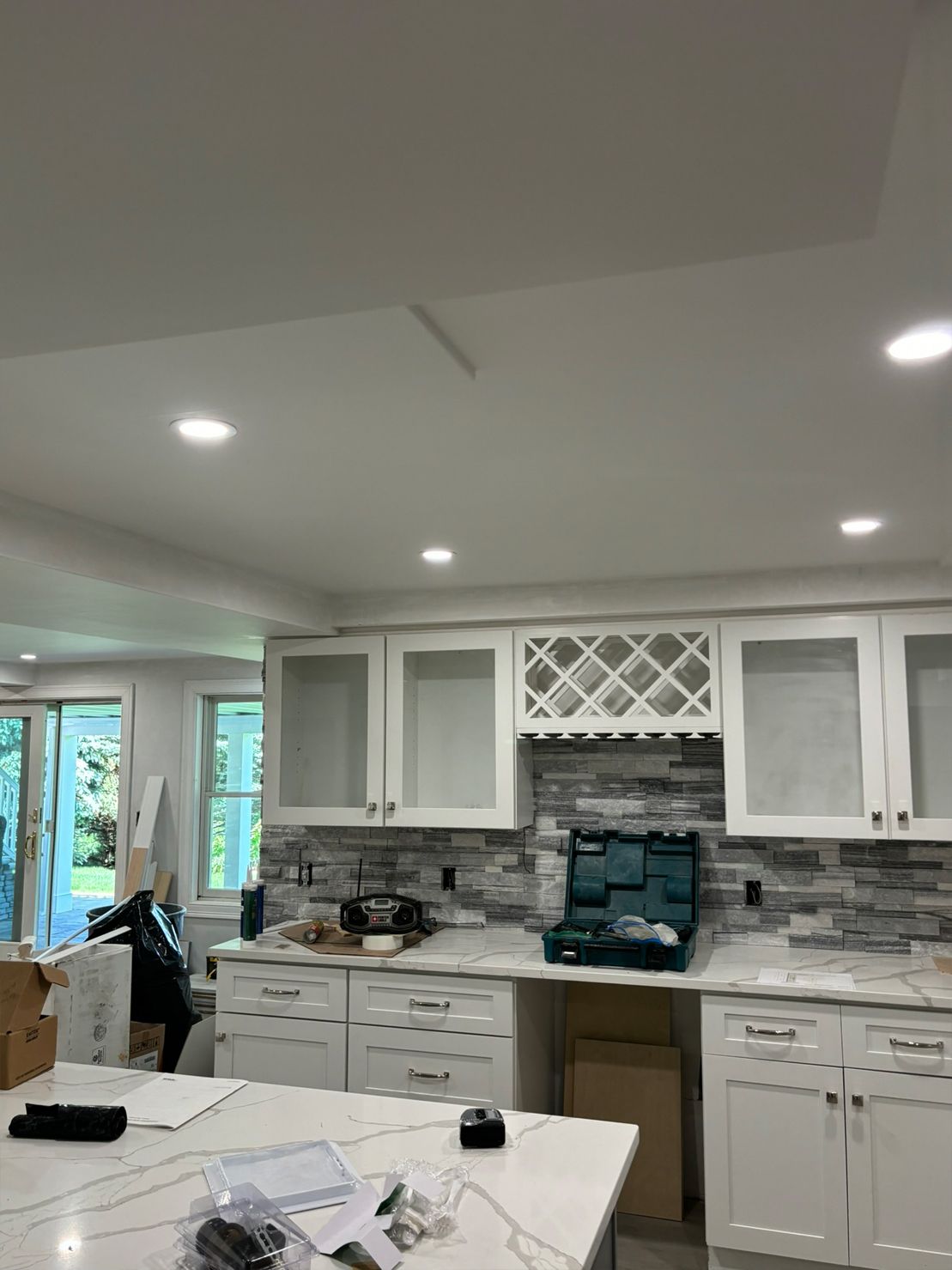 White kitchen cabinets and countertops under construction; stone backsplash, recessed lighting.