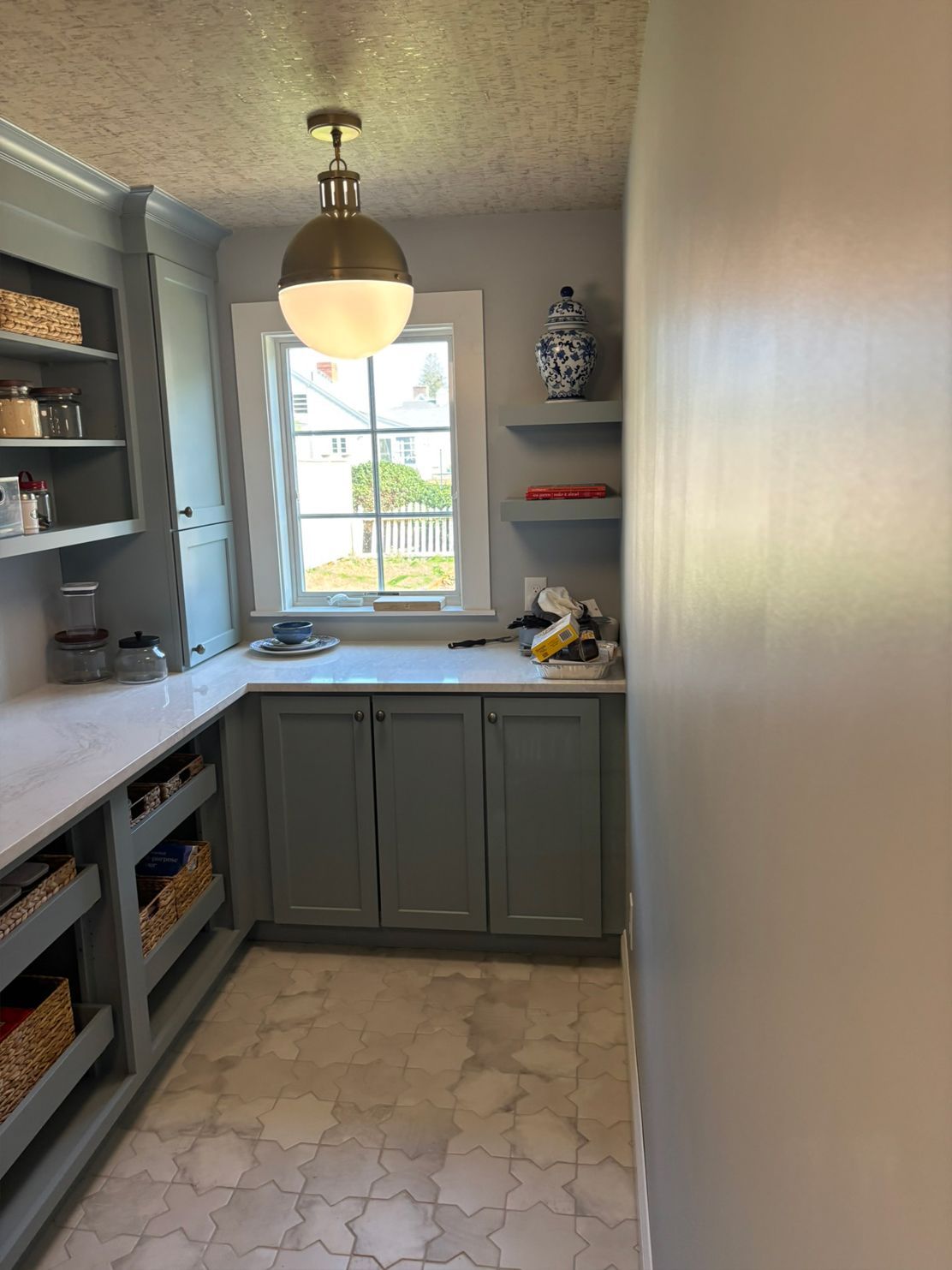 Gray pantry with cabinets, shelves, a window, and a gold and white light fixture.