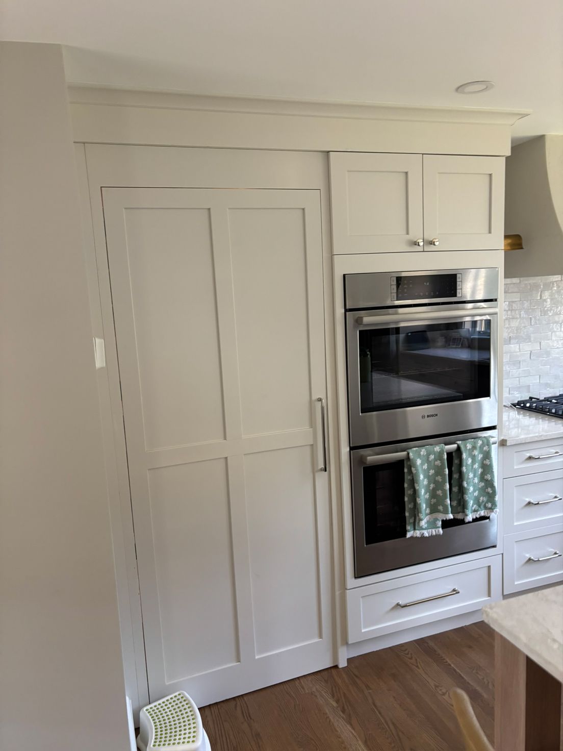 White built-in refrigerator and double oven in a kitchen with white cabinetry and wooden floors.