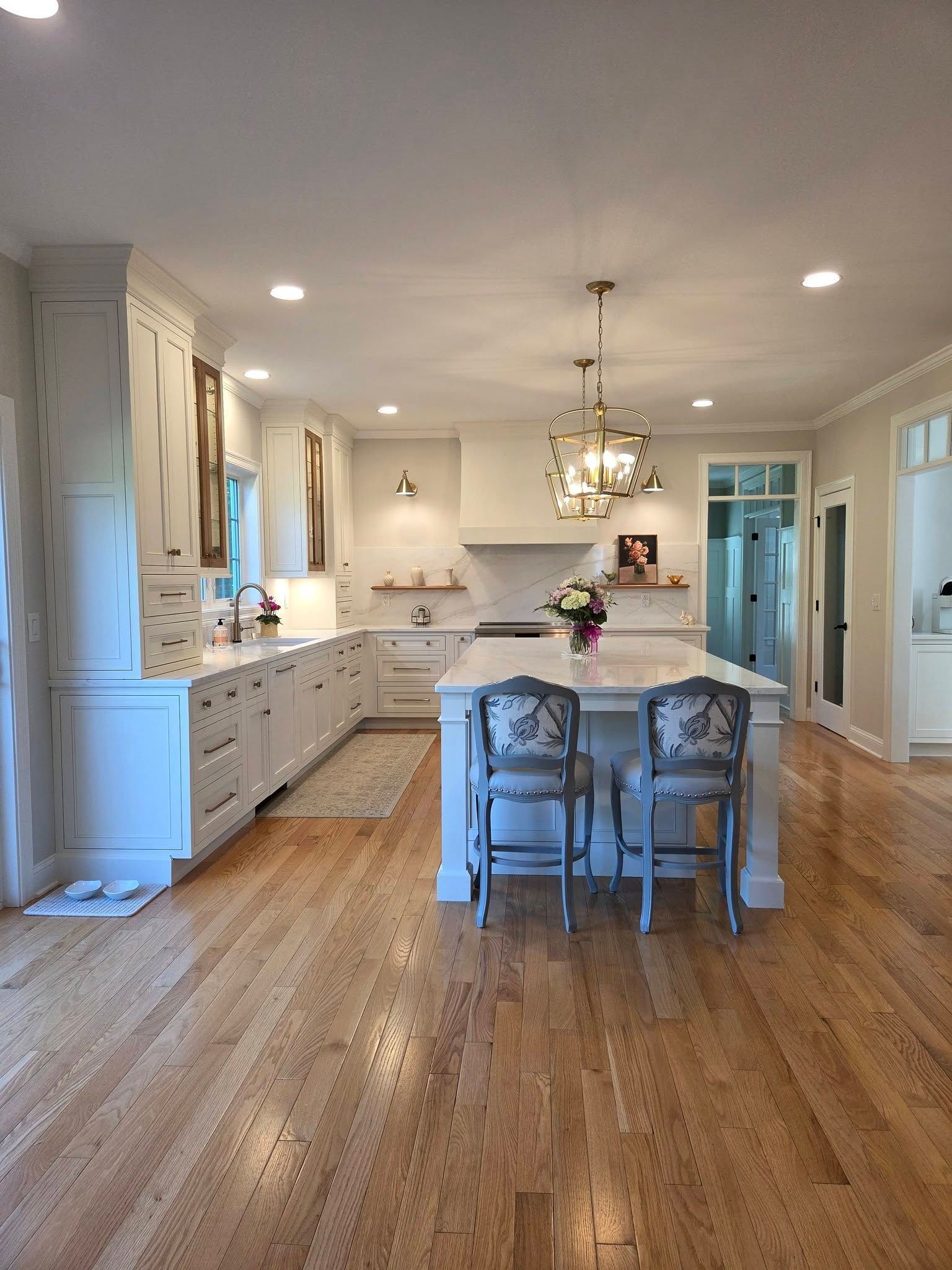 Bright white kitchen with island, pendant lights, and hardwood floors. Two chairs sit at the island.