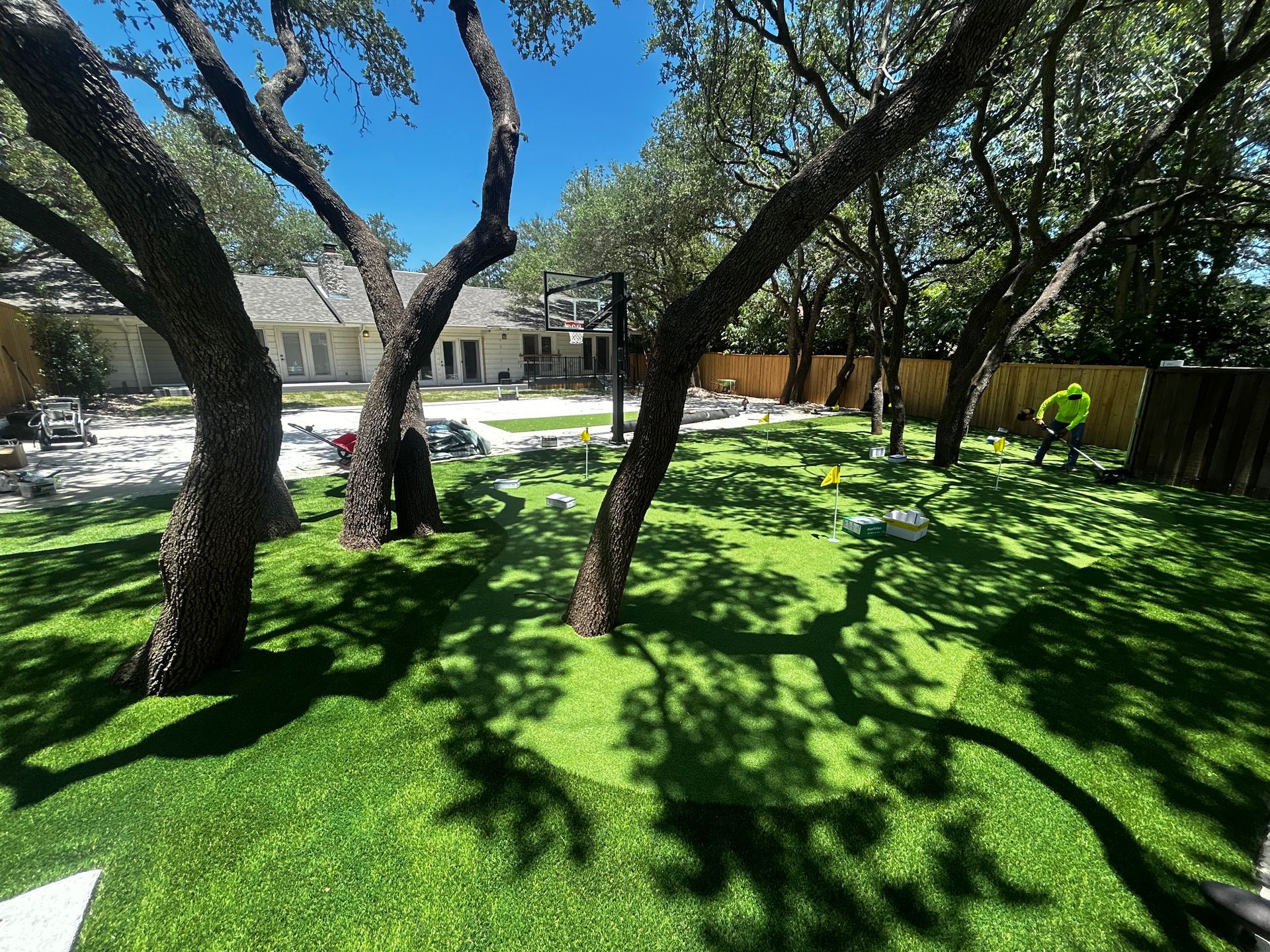Shaded backyard lawn with trees, bright green grass, and a patio beside a wooden fence