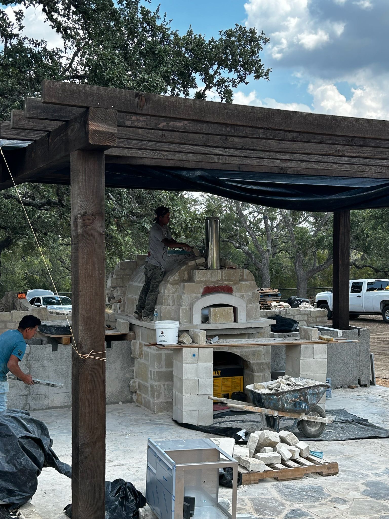 A man is working on a pizza oven under a wooden pergola