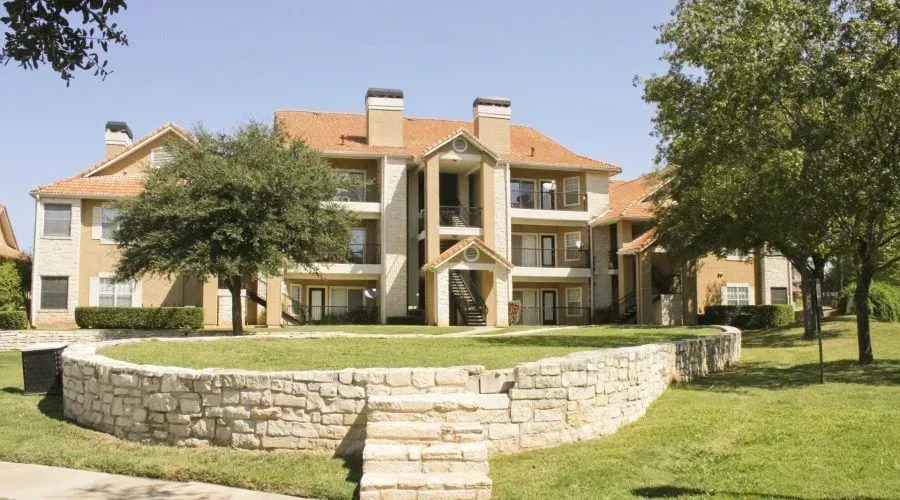 Apartment complex with stone wall, central staircase, and green lawn under trees