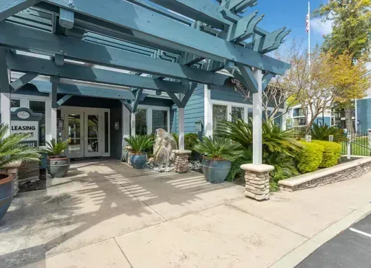 Modern storefront with glass entrance, blue awning, potted plants, and sidewalk seating in bright daylight