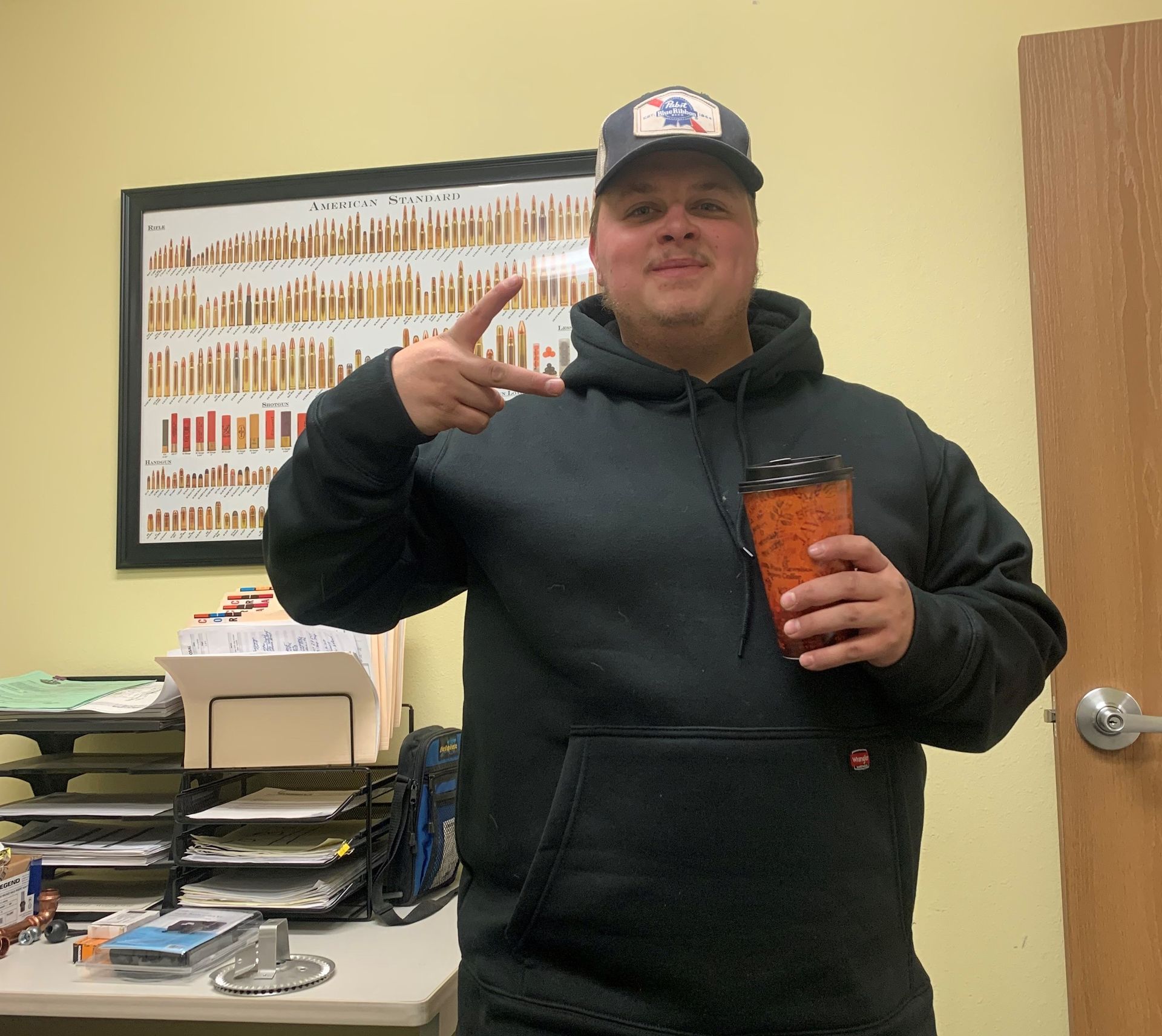 Man in black hoodie and baseball cap holding orange coffee, gesturing peace sign.