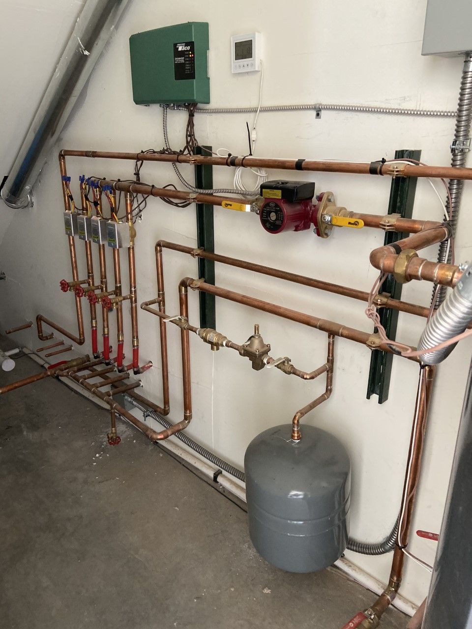 Copper pipe radiant heating system on a white wall. Includes a green control box, a red pump, and a grey expansion tank.