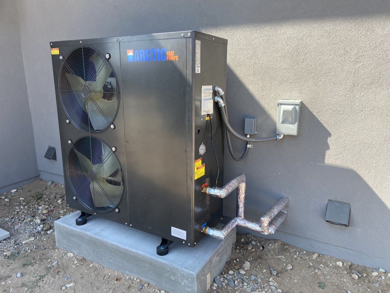 Black heat pump unit mounted on concrete pad, next to a stucco wall.