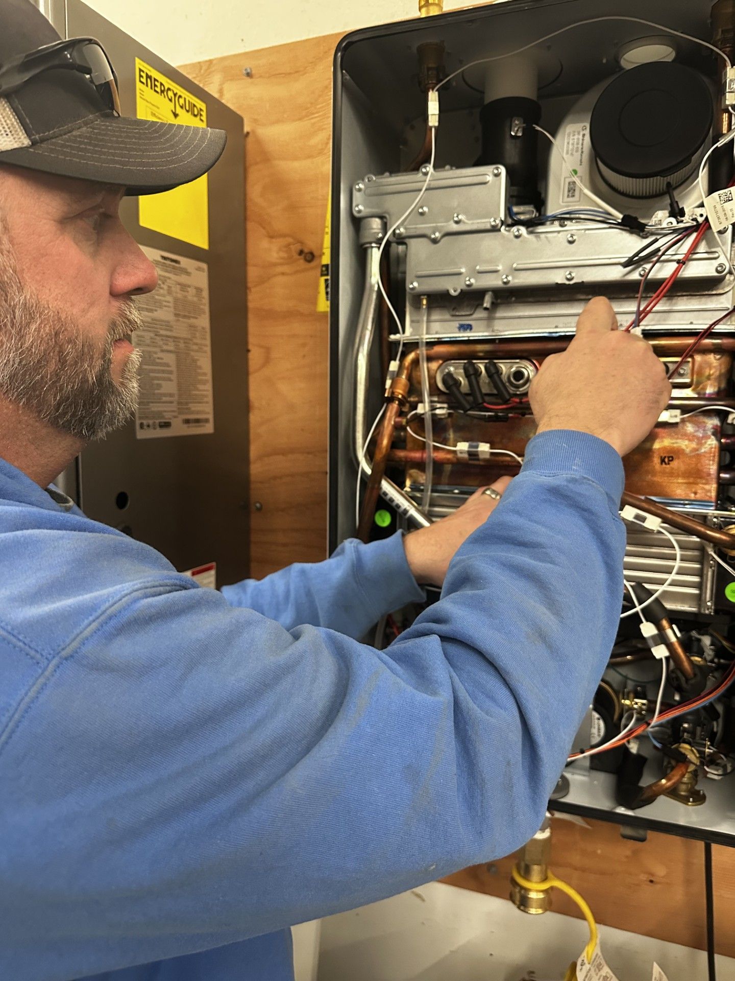 Man in blue sweatshirt works on a water heater, pointing at components with a focused expression.