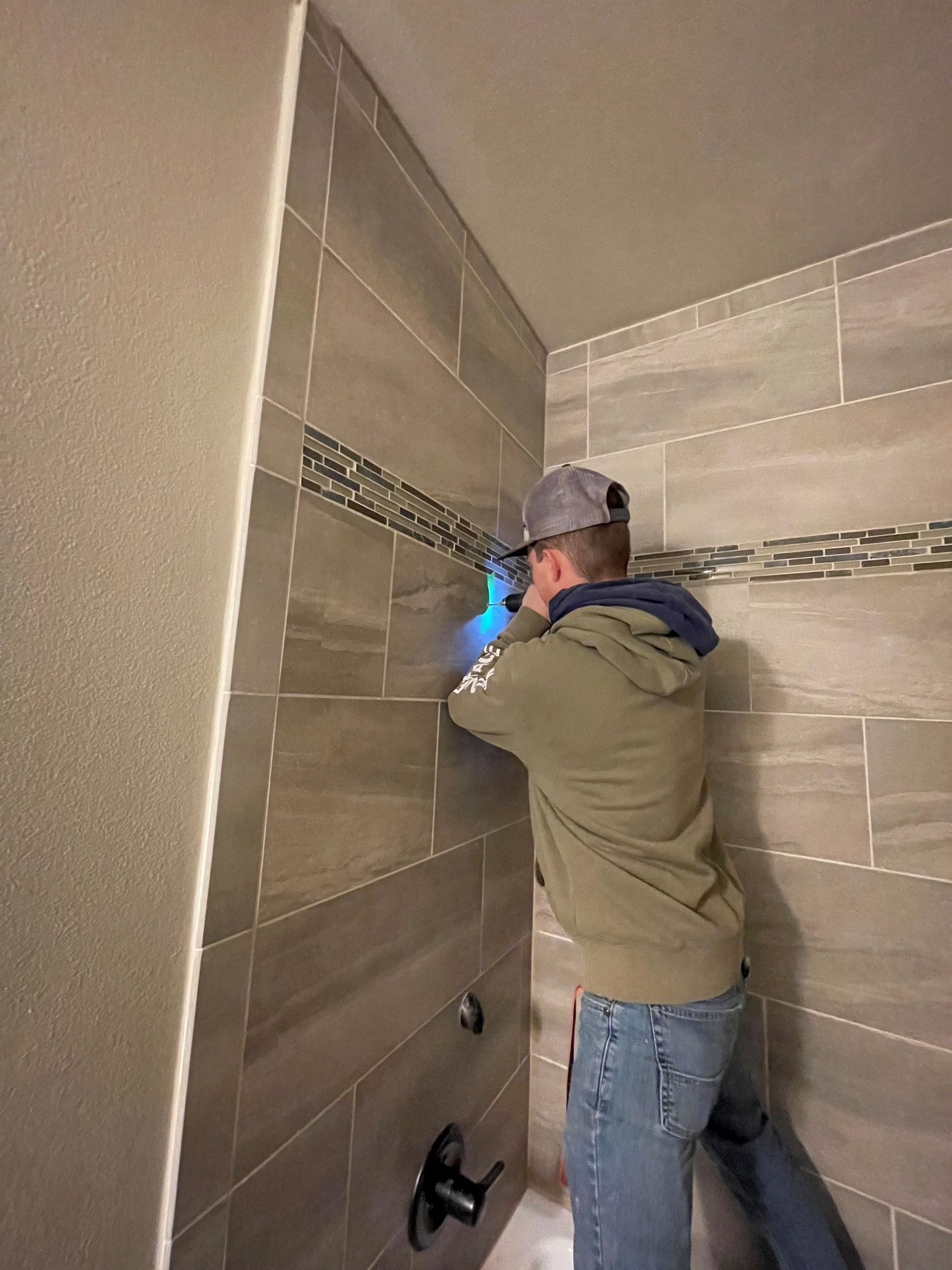 Man in a gray hoodie and jeans working on shower tile; blue light illuminates his hands.