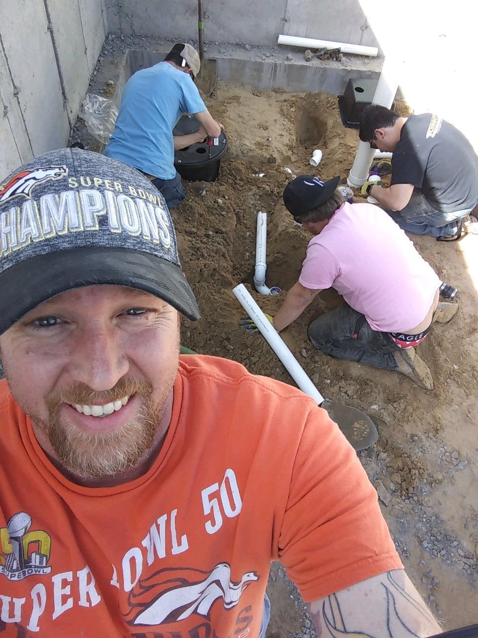 Four construction workers installing pipes in a dirt pit. One smiling wearing orange Broncos gear taking selfie.