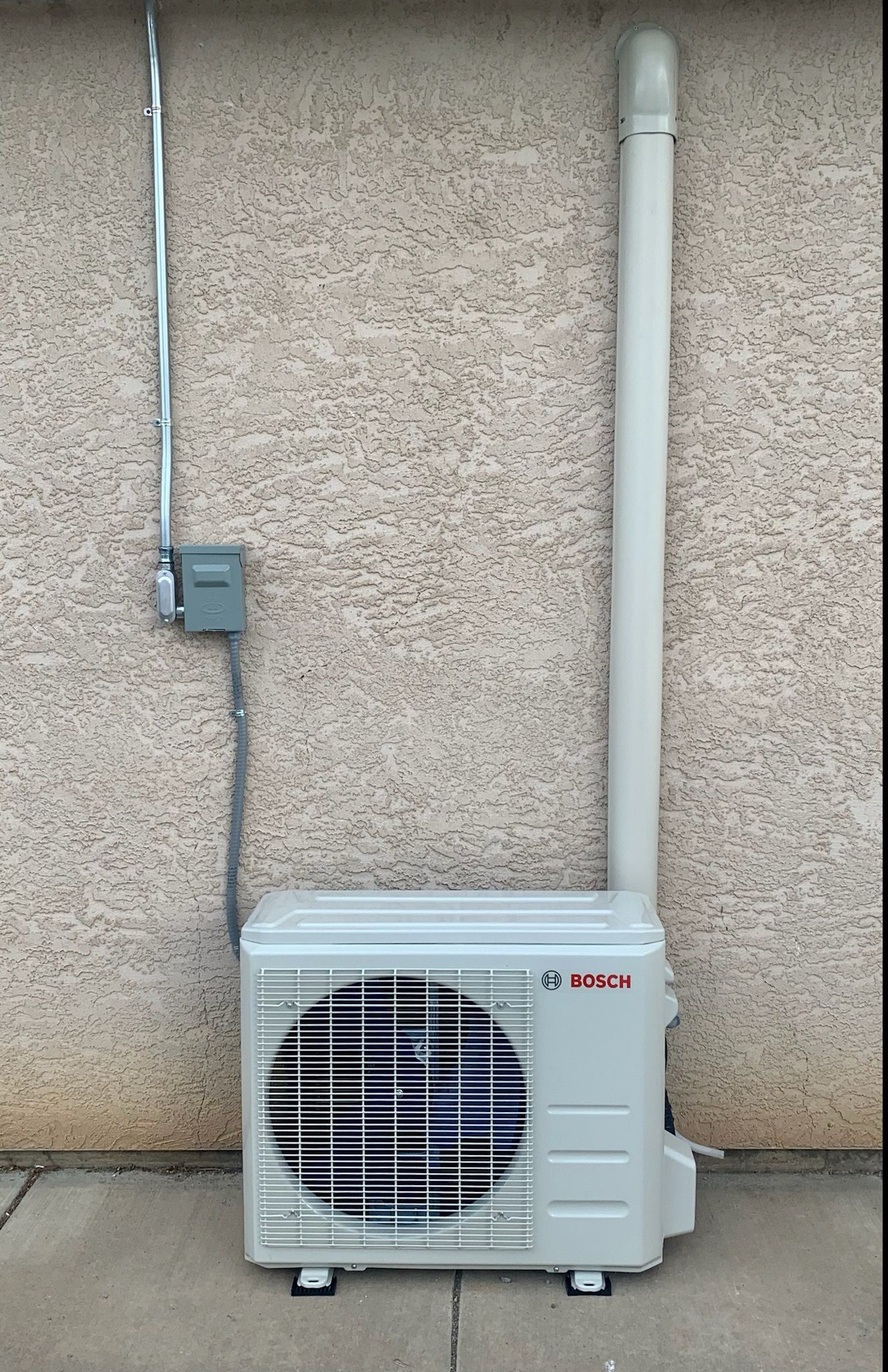 An outdoor air conditioning unit mounted on a beige stucco wall, with a vent pipe extending upward.