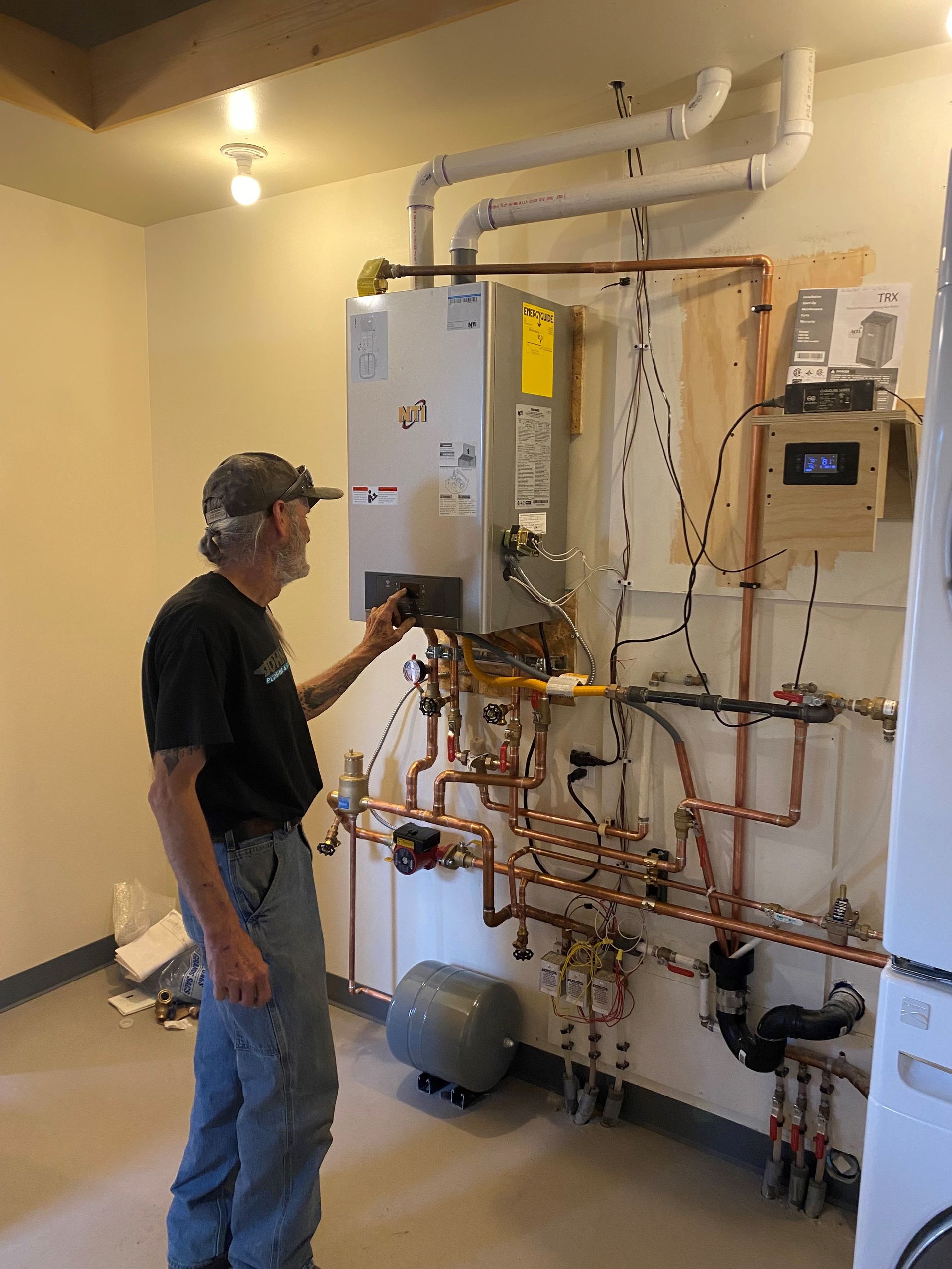 Man inspecting complex plumbing and heating system with copper pipes and tank in a utility room.