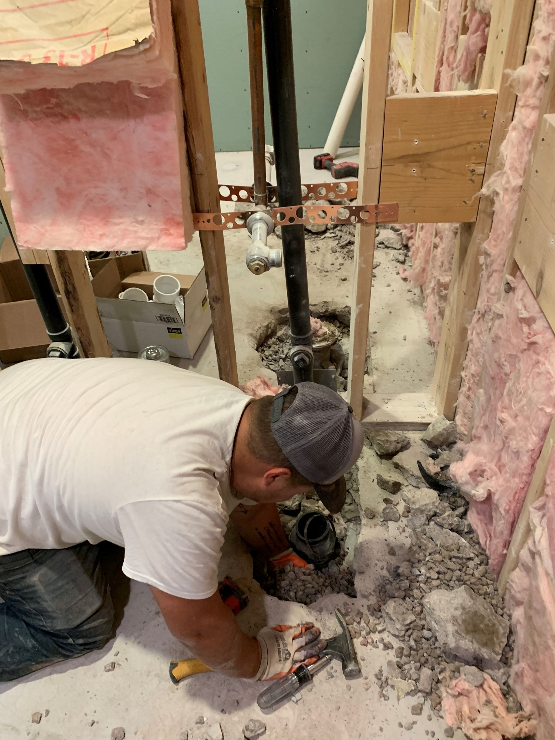 A man in a white shirt works on plumbing, using a hammer and chisel on concrete in a construction area.