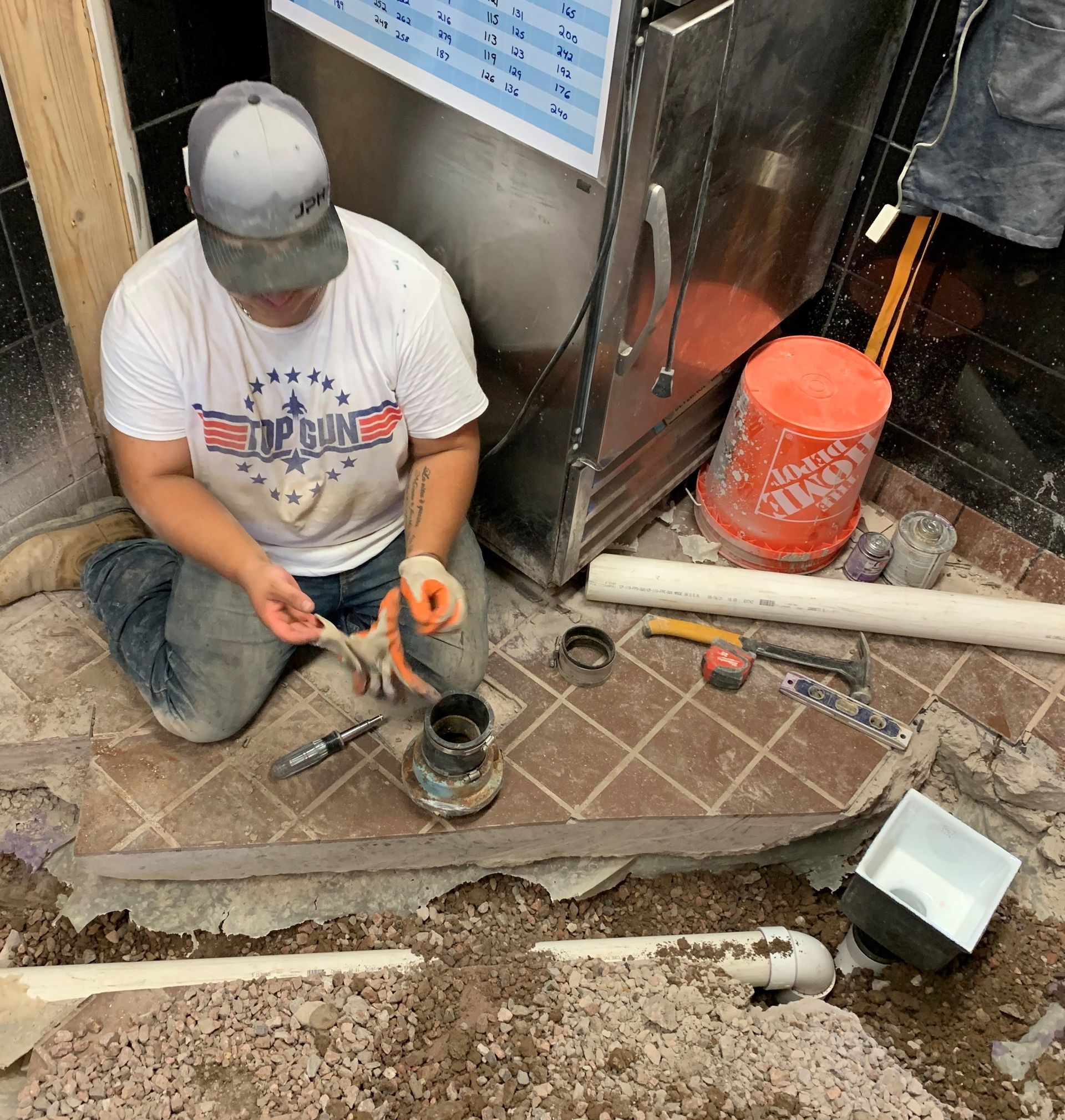 A man wearing a baseball cap and working on plumbing near a red bucket, inside.