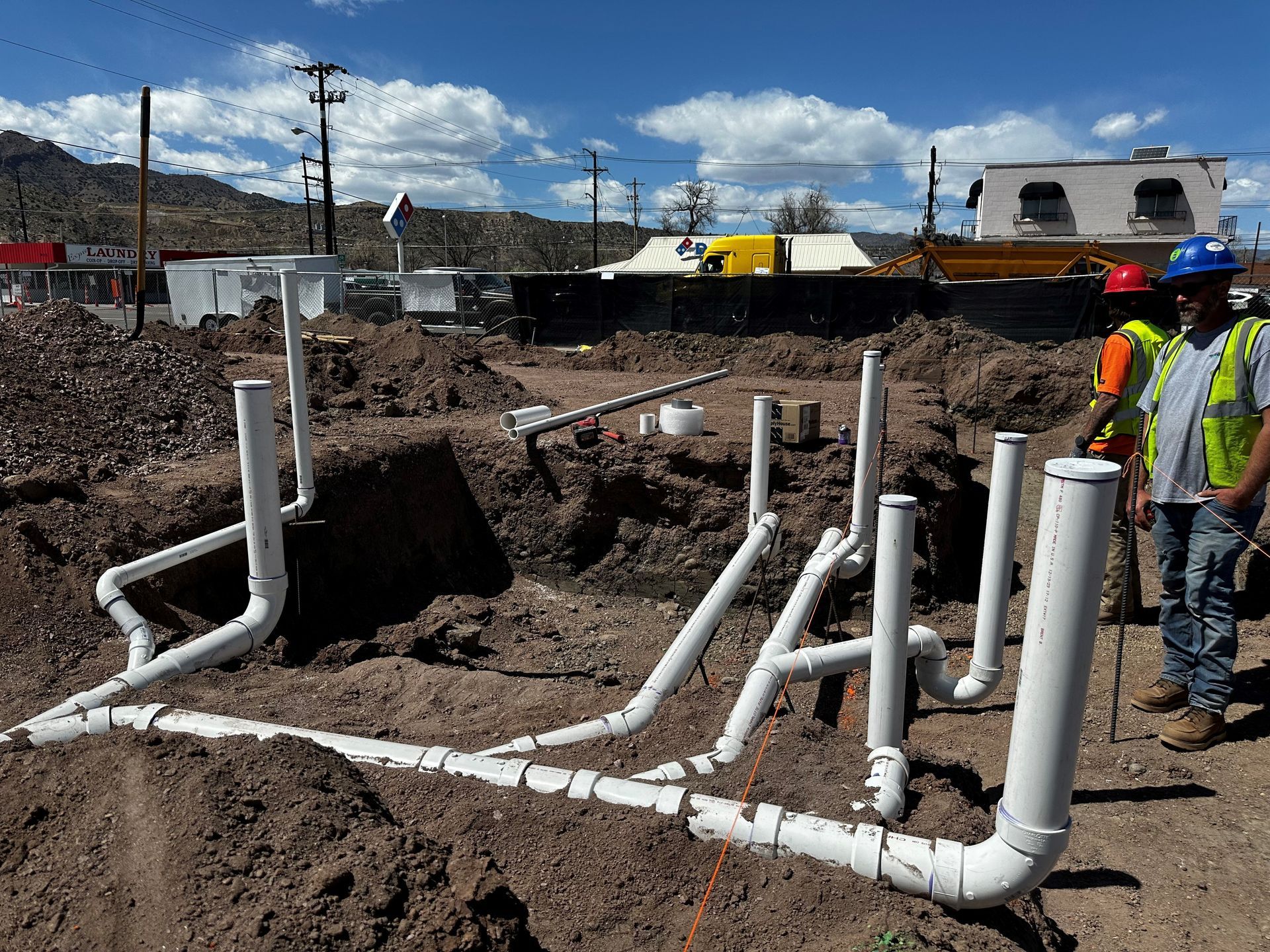 Construction site with white PVC pipes in trenches; two workers in safety vests.