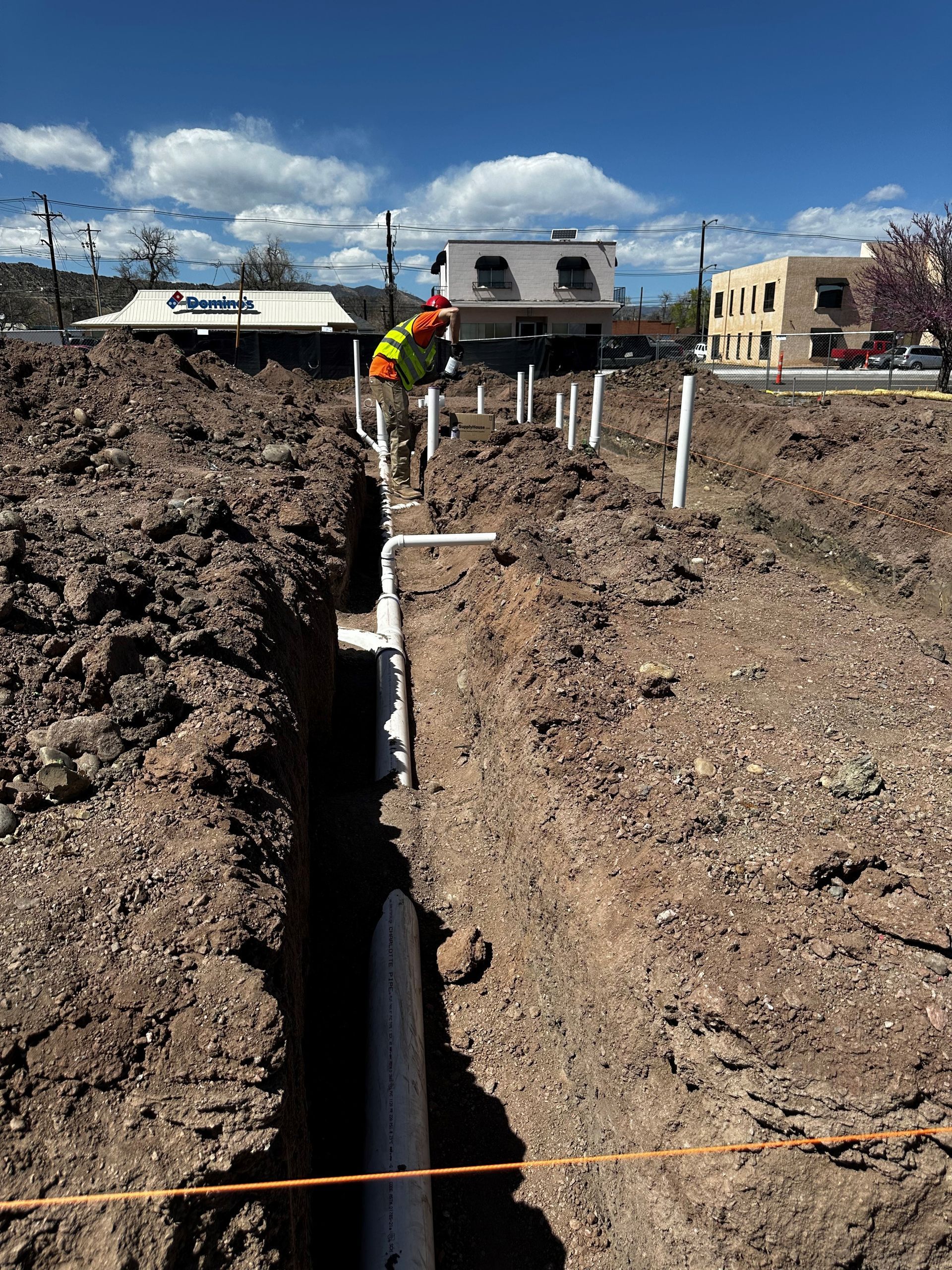 Construction site with trenches and pipes. A worker in an orange vest is working. Buildings and blue sky background.