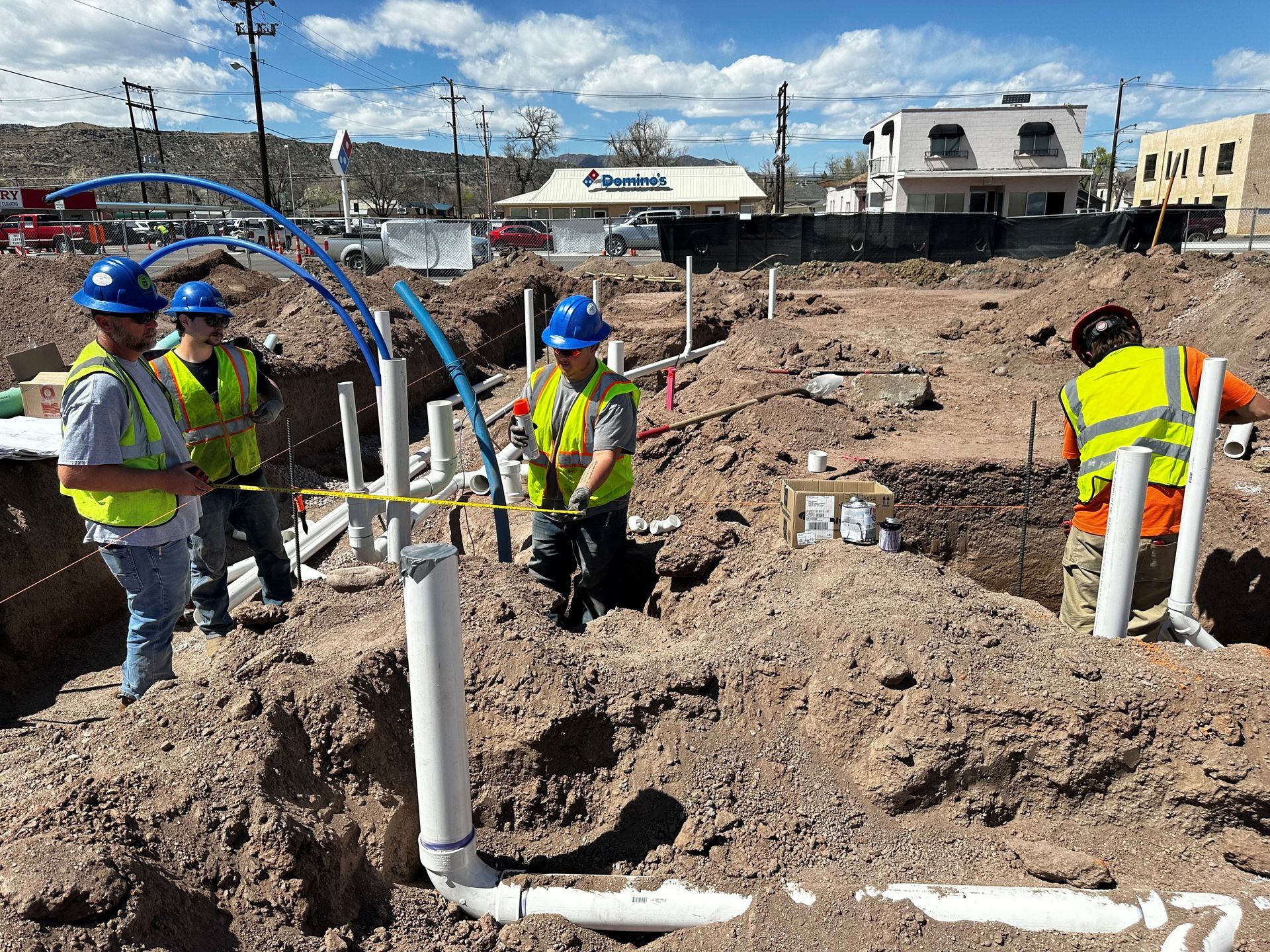 Construction workers installing plumbing in a dirt trench; blue hard hats, yellow vests, outdoors.