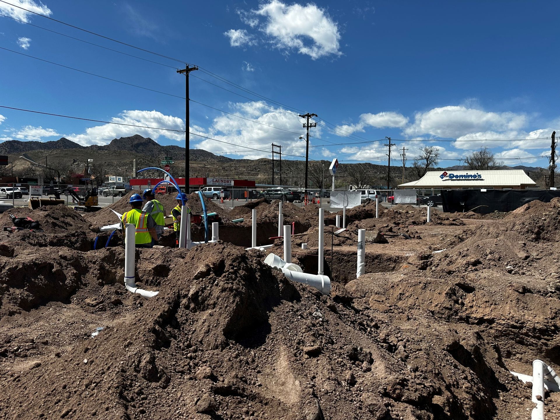 Construction site with exposed pipes; workers in yellow vests; Domino's restaurant in the background.