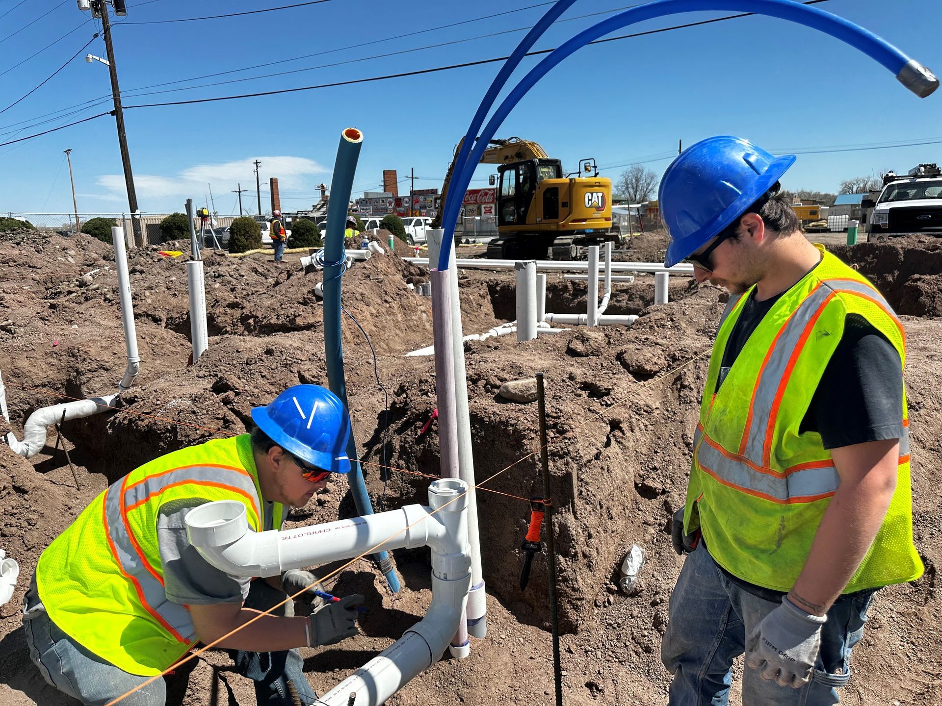 Two construction workers in hard hats and vests connecting pipes in an excavation site.