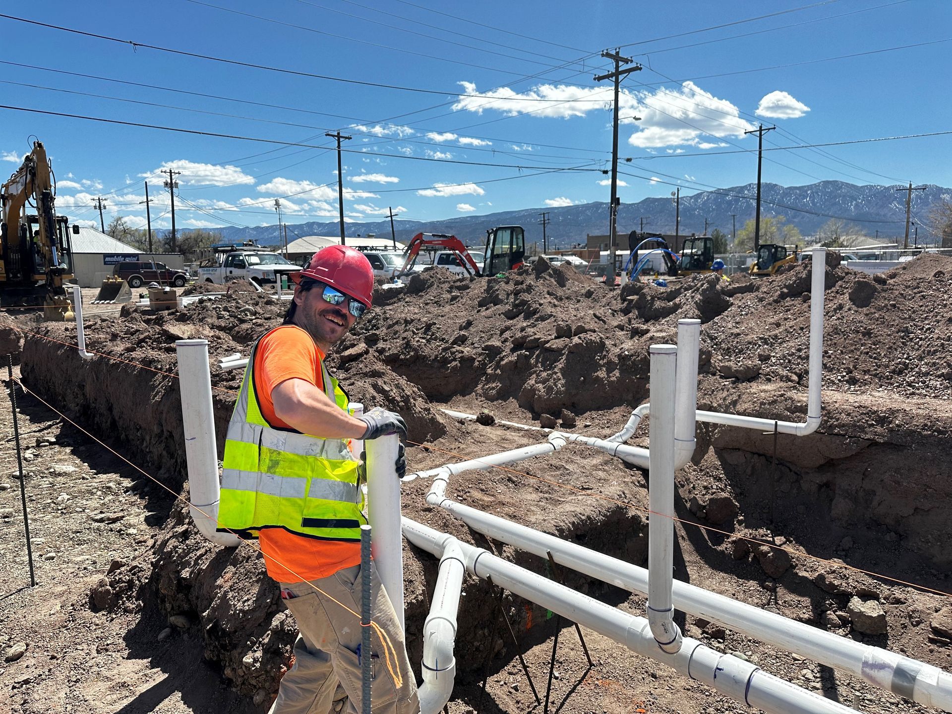 Construction worker in a hard hat and vest smiles, working on white pipes in a dirt trench; outdoor construction site.