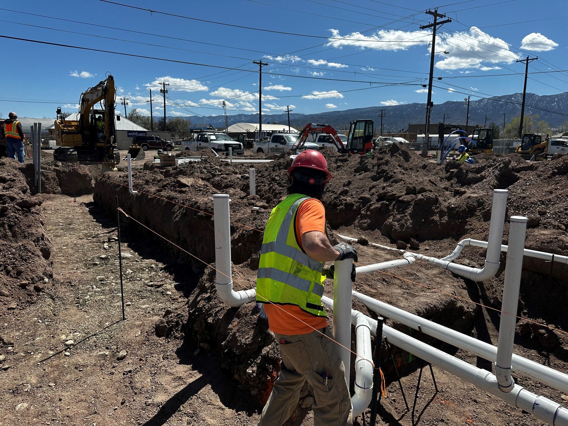 Construction worker in hard hat and safety vest installs PVC pipes in a trench. Sunny day, outdoor site.