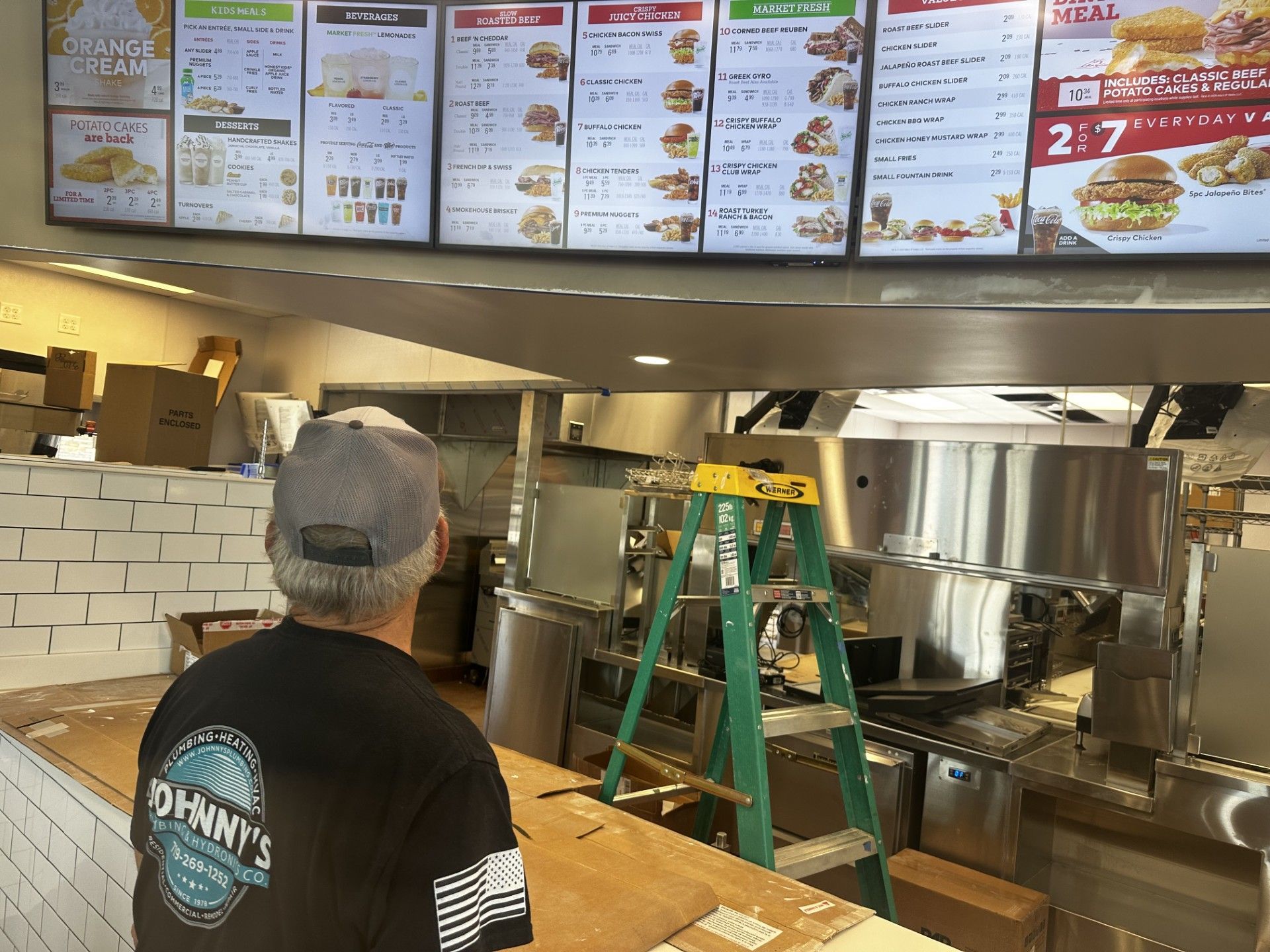 A man in a grey hat looks at digital menu boards in a partially constructed restaurant kitchen.