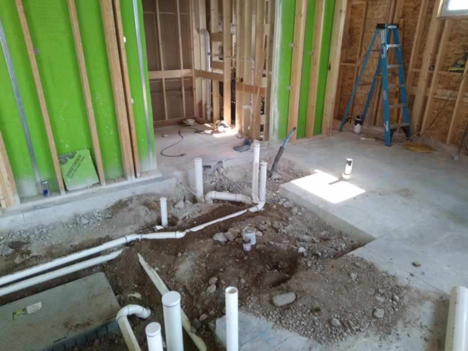 Interior of a building under construction, plumbing pipes in the concrete floor. Stud walls are exposed, green insulation visible.