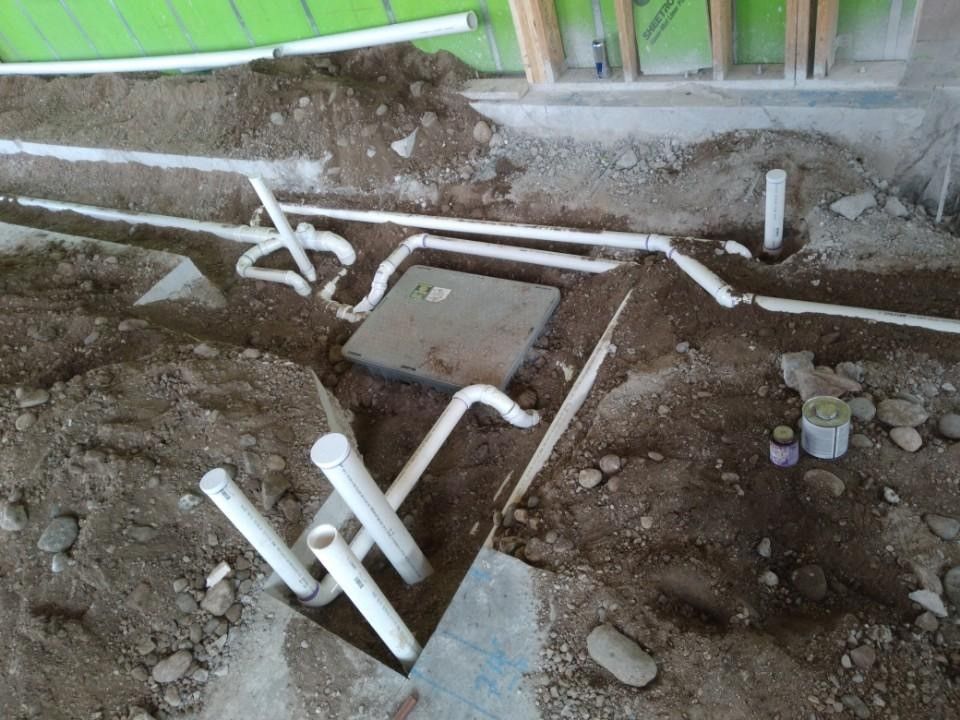 White PVC pipes and electrical box in a dirt trench, preparing for plumbing and electrical work in a construction setting.