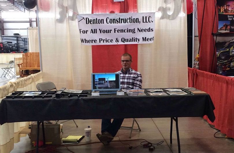A person sits at a black-covered table with a laptop, under a Deaton Construction sign at a trade show booth.