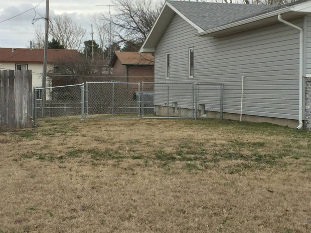 A side view of a gray-sided house with a fenced-in yard and dormant, patchy grass on an overcast day.