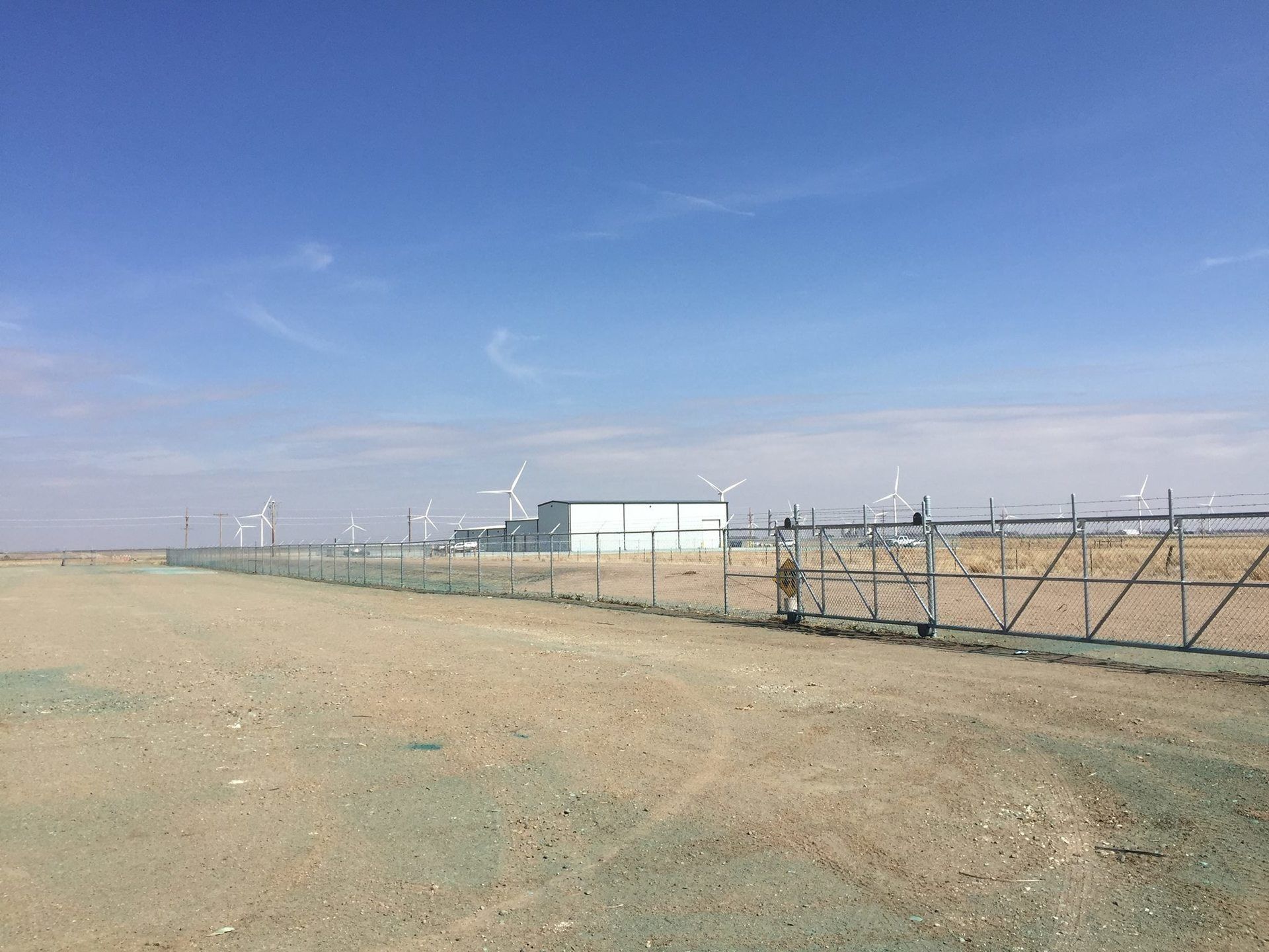 A fenced-in industrial site under a clear blue sky, featuring a modern building surrounded by distant wind turbines.