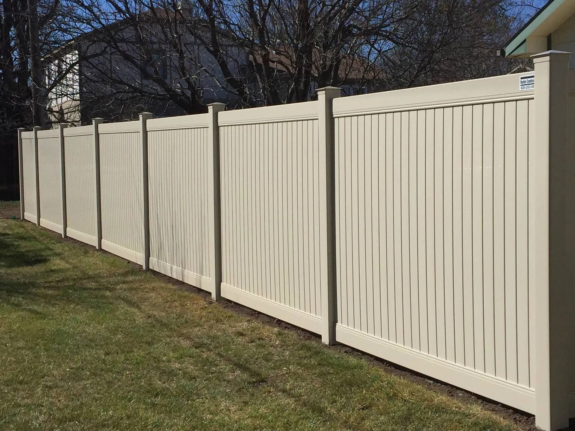 A tan vinyl privacy fence stretches across a green lawn on a sunny day.