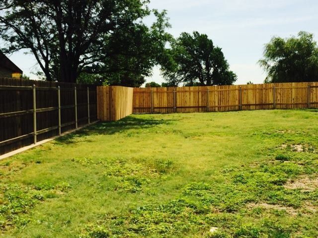 A grassy backyard enclosed by a wooden fence with mature trees in the background under a clear sky.
