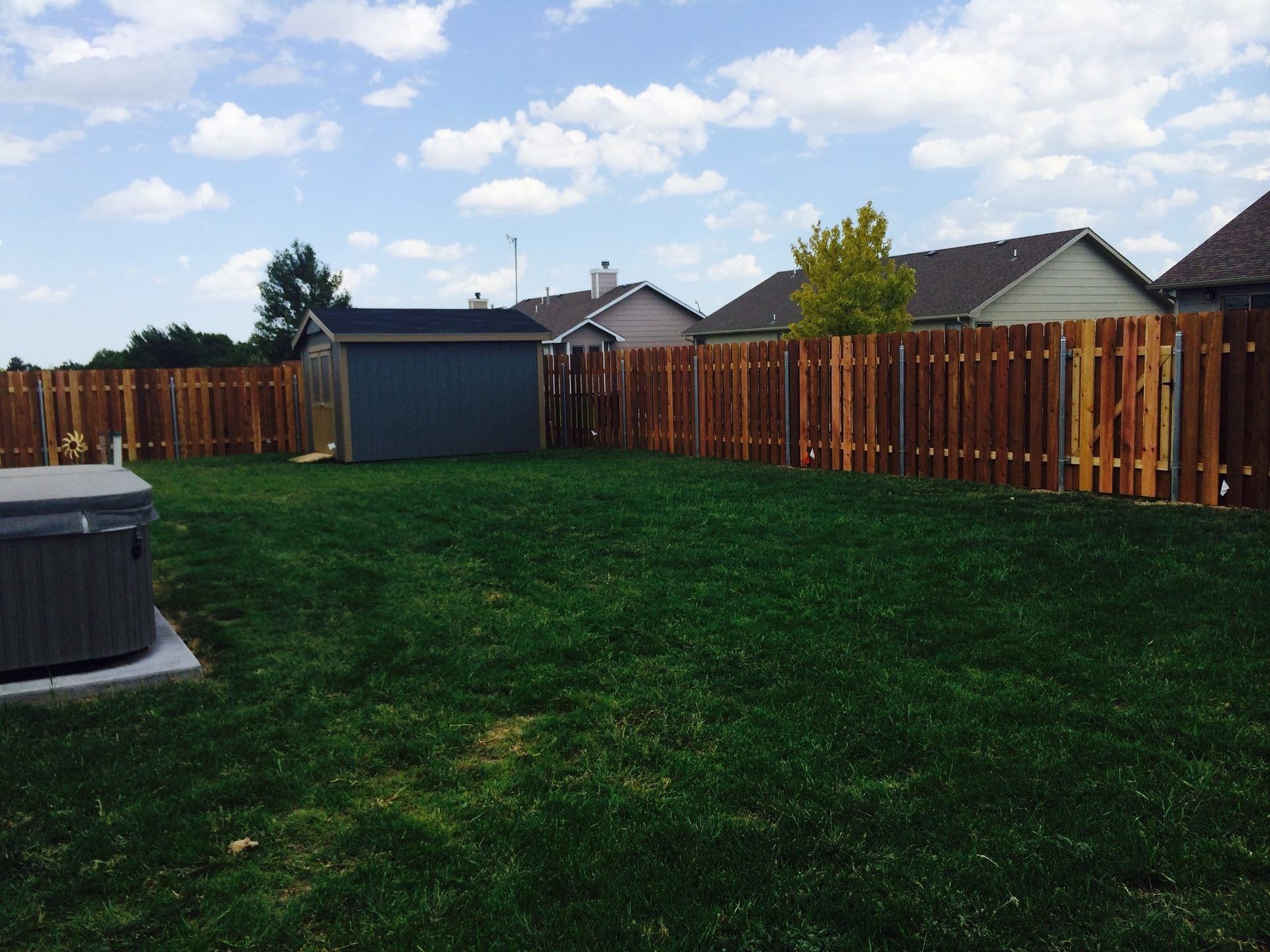 A backyard with a grassy lawn, a small grey shed, a hot tub, and a tall wooden fence under a blue sky with clouds.