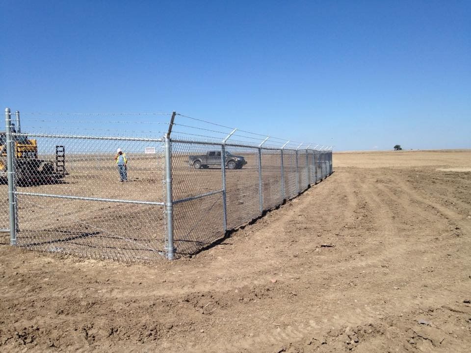 A chain-link fence with barbed wire tops a dirt construction site under a clear blue sky, with a worker and truck nearby.
