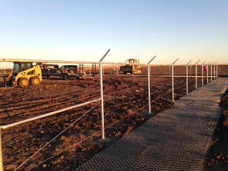 Construction crew installs chain-link fence posts on a muddy field with heavy machinery in the background at sunset.