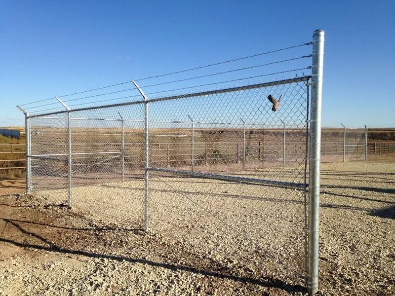 A tall chain-link security fence with barbed wire on top stands on a gravel patch under a clear blue sky.