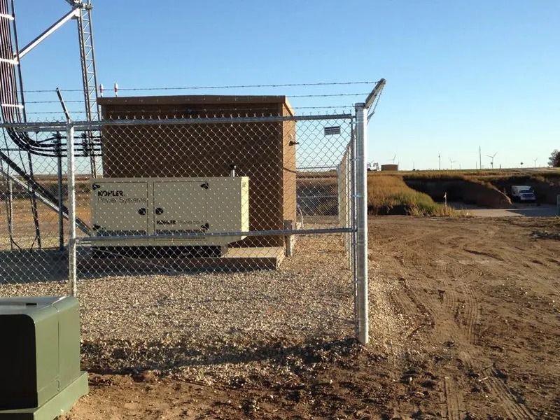 A beige generator sits inside a fenced compound next to a small utility building on a gravel lot under a clear blue sky.