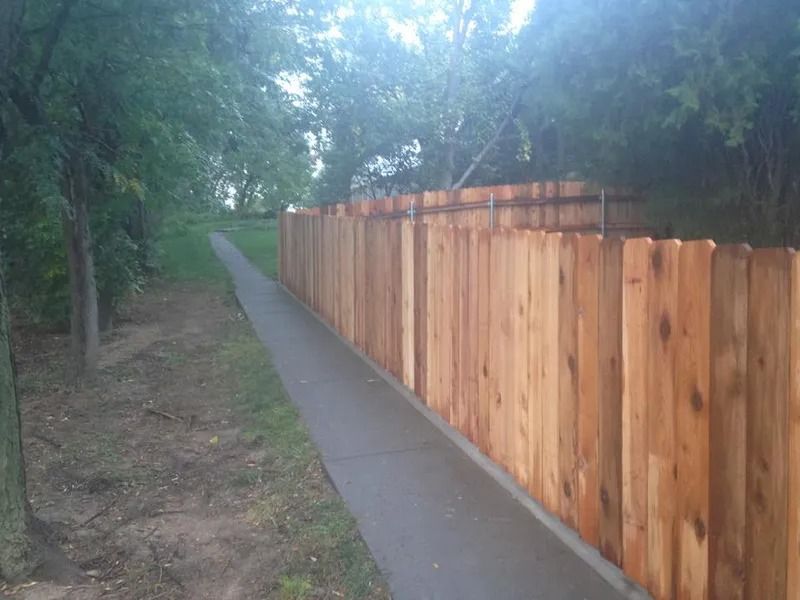 A new, light-colored wooden fence runs alongside a paved sidewalk next to a grassy area with trees.