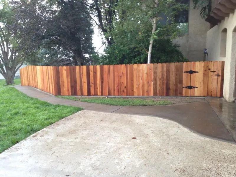 A newly installed cedar wood fence curves along a paved driveway next to a grassy yard and a house.
