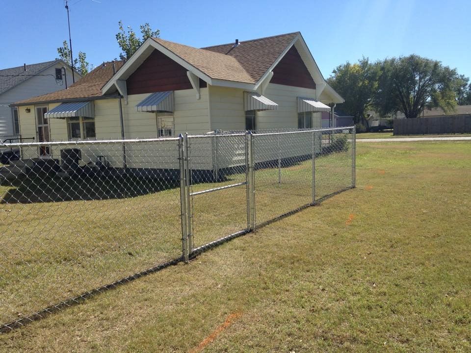 A yellow, single-story house with dark red gables is partially enclosed by a chain-link fence in a grassy yard.
