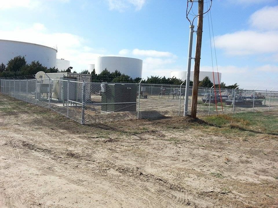 A chain-link fence encloses industrial equipment and several large, white storage tanks under a partly cloudy sky.