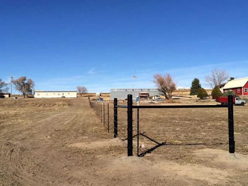 A new black metal fence and gate installed on a rural property with a dirt field, bare trees, and buildings in the distance.