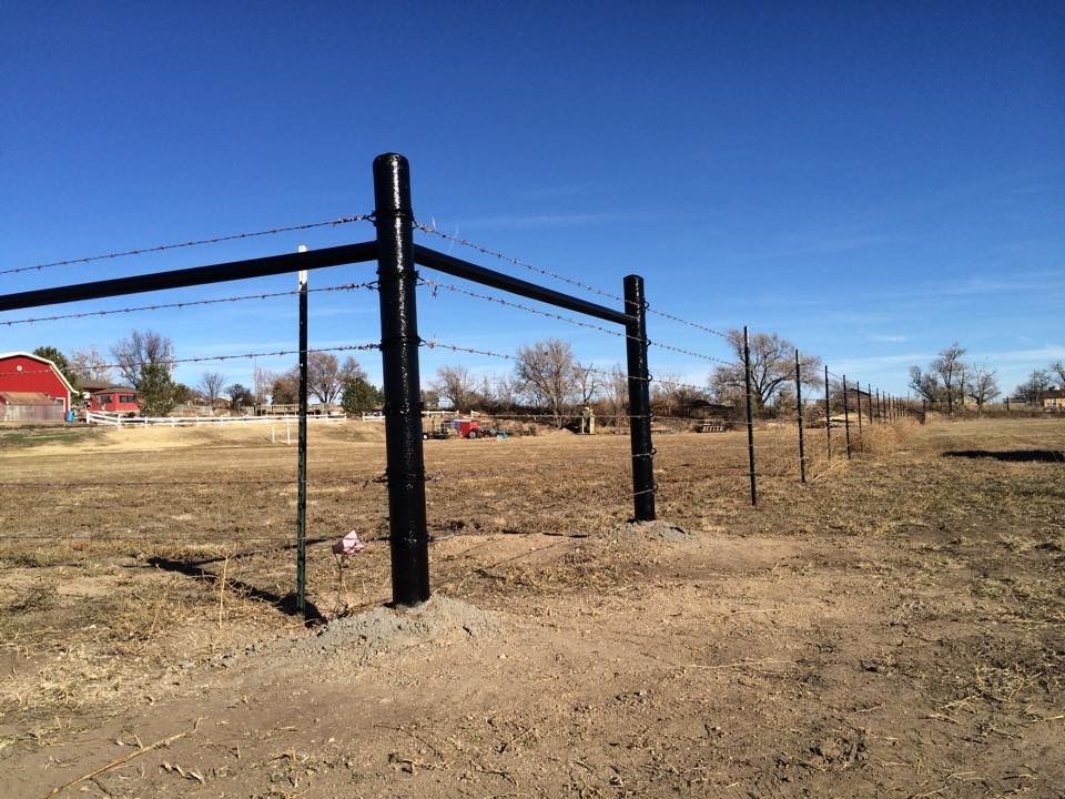 A wire fence with black posts runs across a dry, brown field under a clear blue sky.