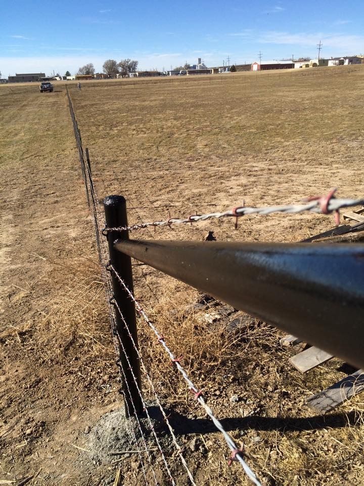 A black metal fence post stands in a dry, open field with barbed wire lines extending into the distance under a blue sky.